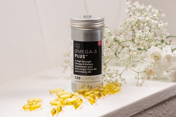 Container of Og Omega-3 Plus capsules with some capsules displayed in front of it and flowers behind 